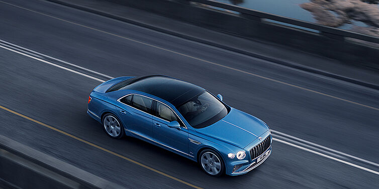 Bentley Lisboa New Bentley Flying Spur Azure sedan in Light windsor blue paint by Mulliner, with 22 inch Azure wheels seen from overhead driving dynamically on a country road