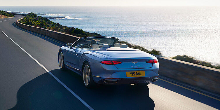 Bentley Lisboa New Bentley Continental GTC Azure convertible rear view in Light Windsor blue paint with 22 inch ten spoke wheels driving dynamically on an ocean road in the sunshine